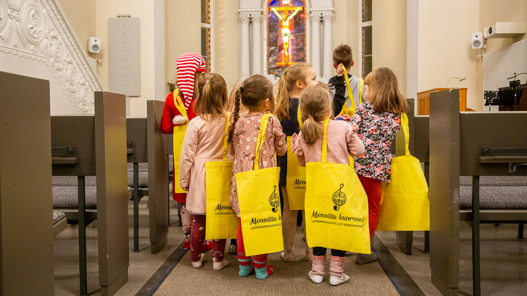 Lapset kävelevät Lpr kirkon  käytävää pitkin. Selässä keltaiset kangaskassit tekstillä 