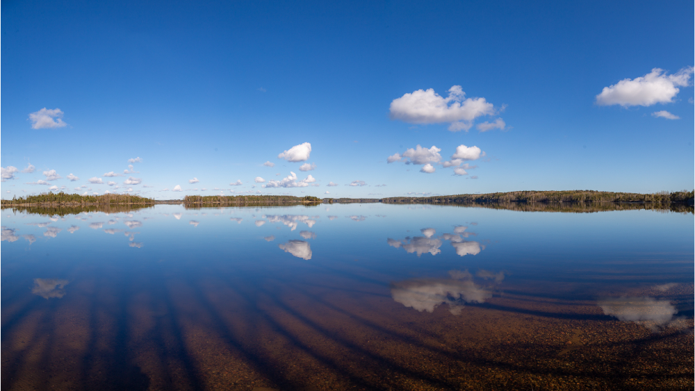 Tyyni Saimaa, jossa horisontissa saaria ja valkoiset pilvet heijastuvat veteen.