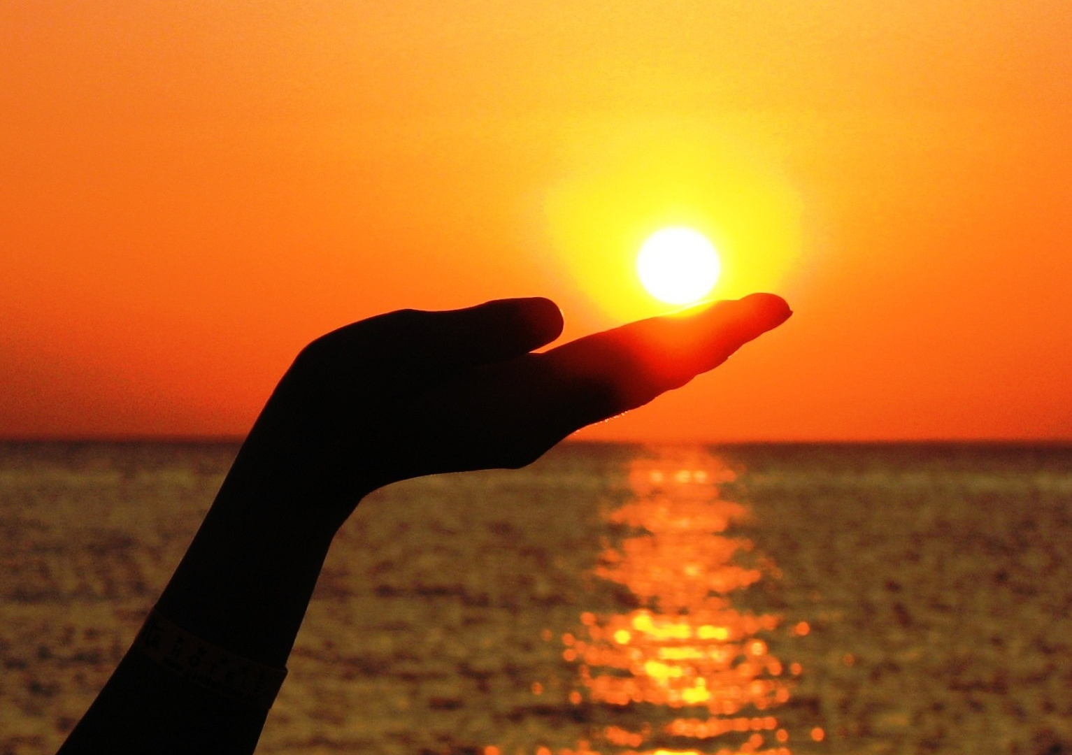 Ilta-aurinko ja meri taustalla, etualalla käsi pitelemässä aurinkoa.