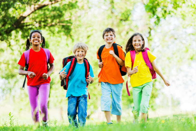 Kouluikäisiä poikia ja tyttöjä värikkäissä paidoissa, reput selässä kävelemässä nurmikolla puistossa.