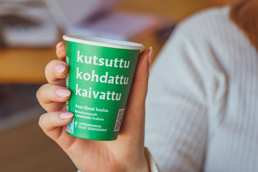 Nainen pitää kädessään vihreää seurakuntavaalit mukia, jossa teksti kutsuttu-kohdattu-kaivattu.